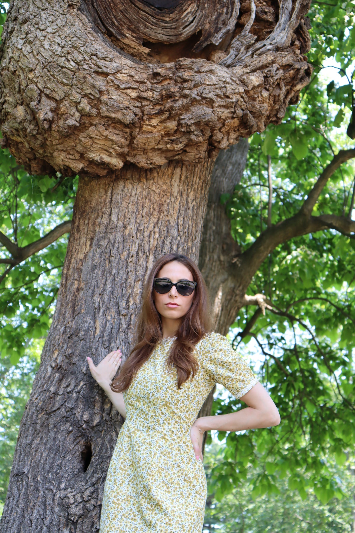Close up photo of Model in short yellow floral dress with short sleeves and white daisy trim leaning on a tree.