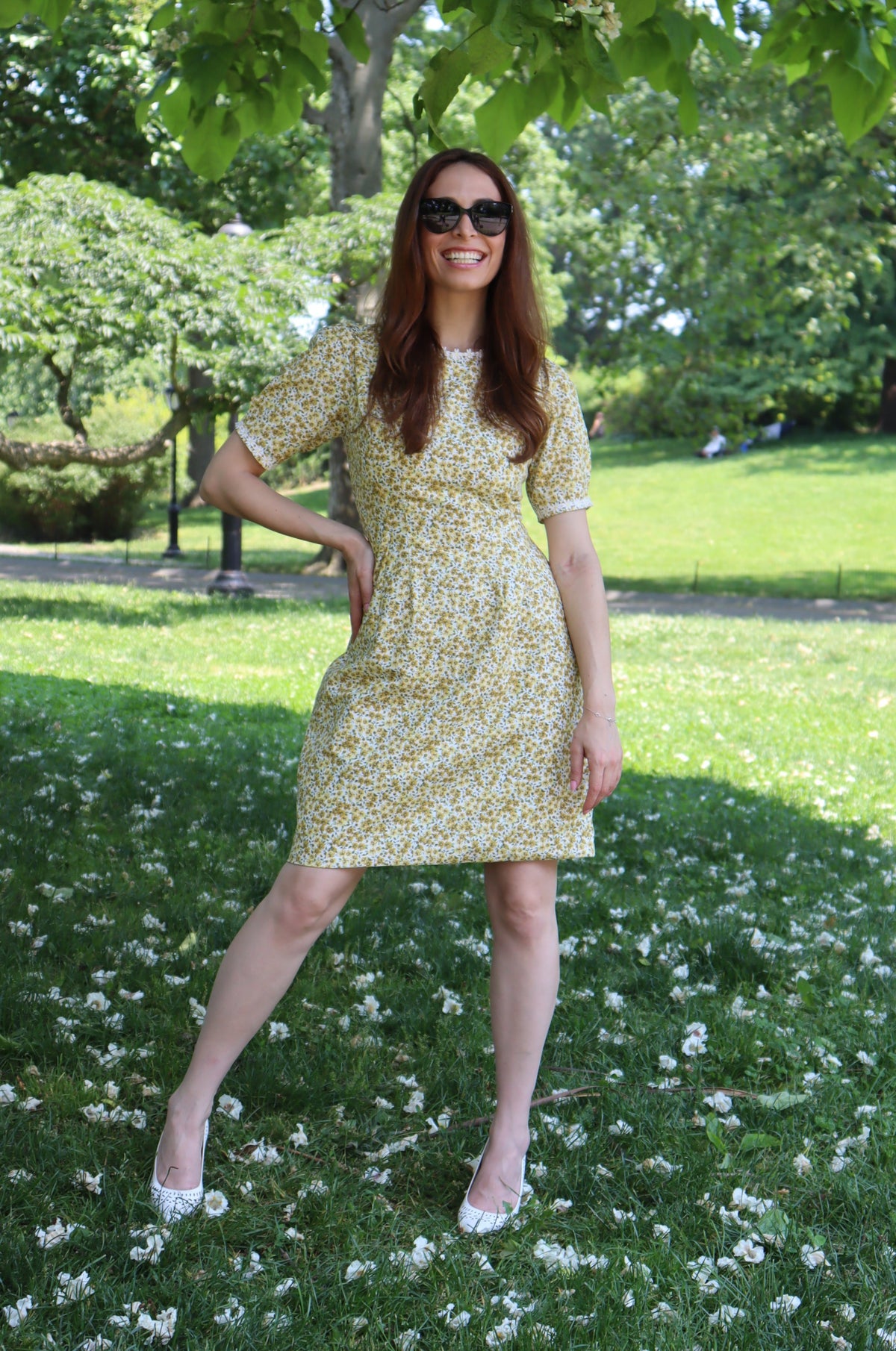 Model in short yellow floral dress with short sleeves and white daisy trim with trees in the background.