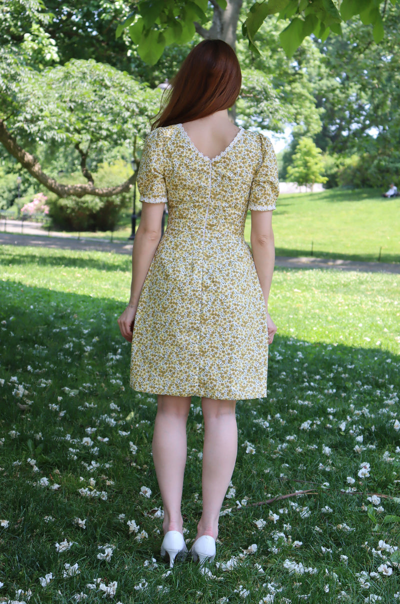 Photo of the back of a model in short yellow floral dress with short sleeves and white daisy trim with a back zipper.