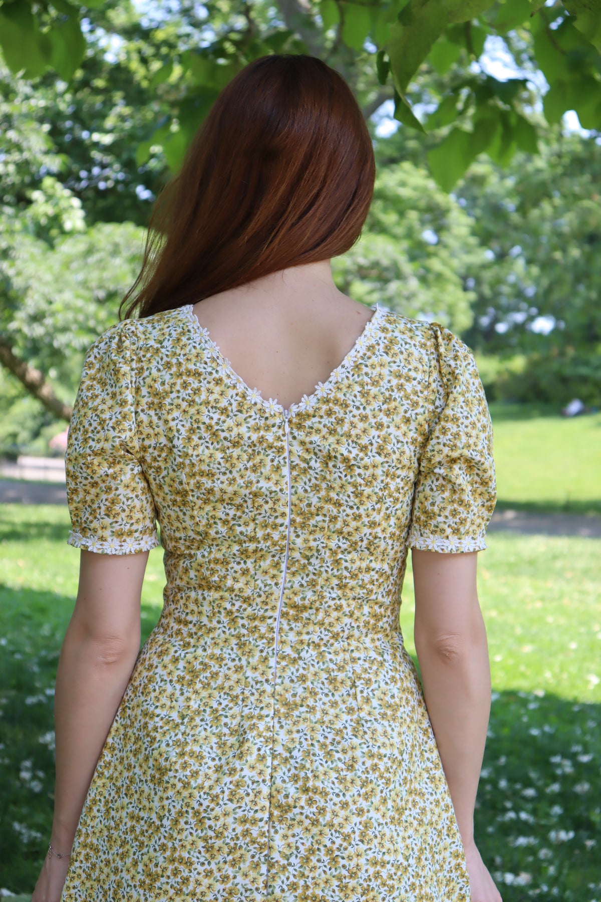 Photo of a model showing the detail of the back neckline of a short yellow floral dress with short sleeves and white daisy trim .
