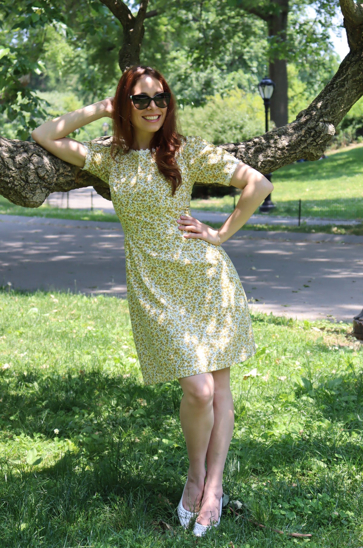 Model in short yellow floral dress with short sleeves and white daisy trim leaning on a tree.