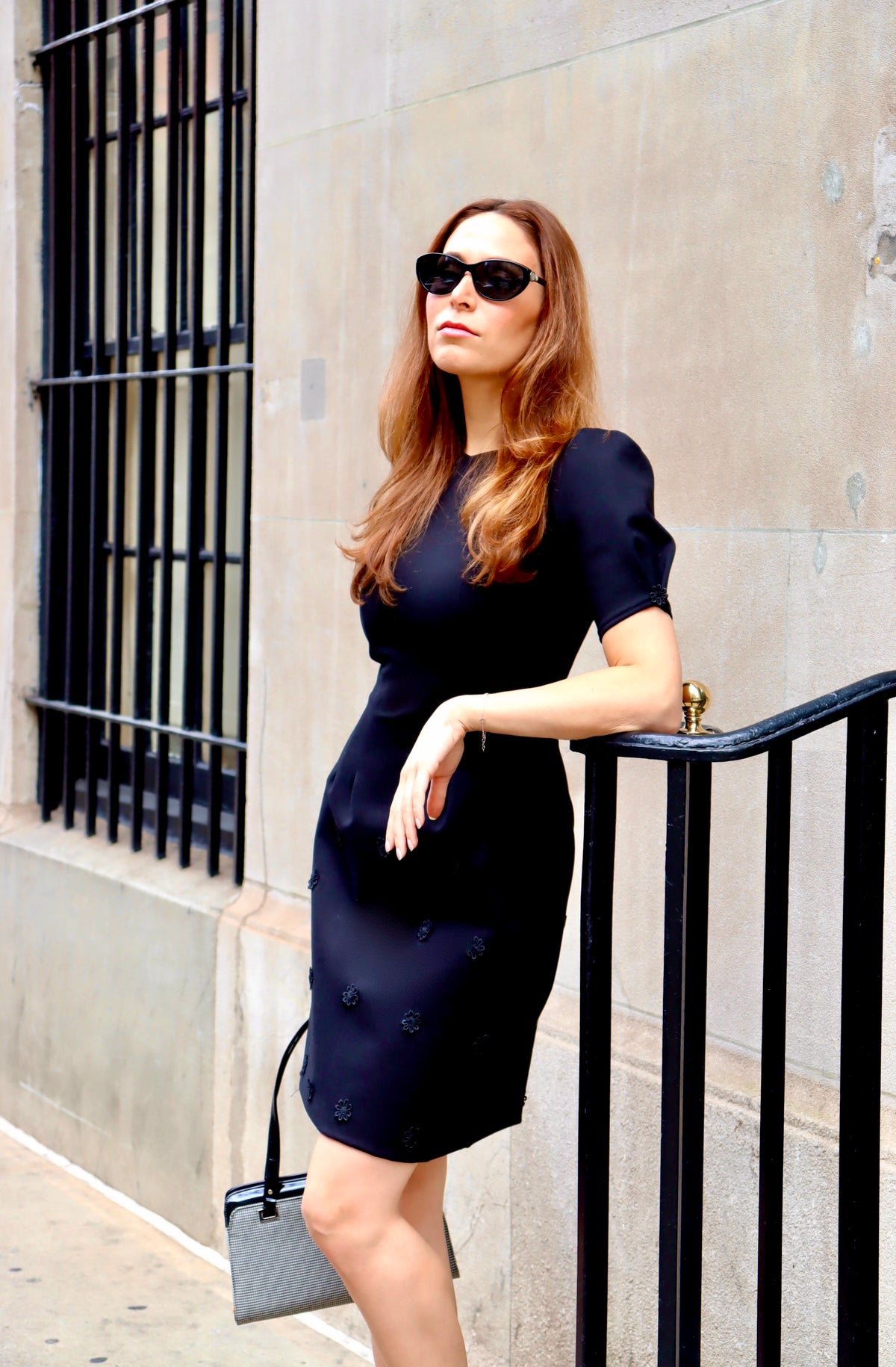 Model leaning on a railing, and stunning in our Fall in Love Floral Appliquéd Black Ponte Dress, sporting sunglasses and a classic bag and heel combo