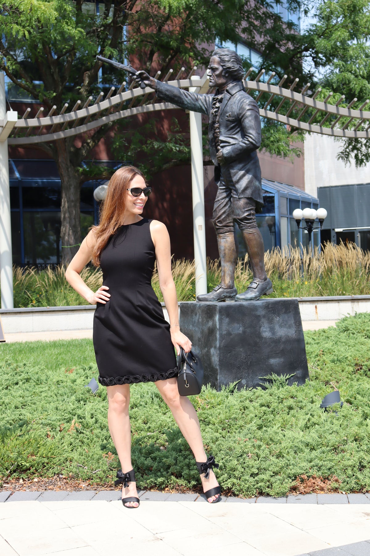 Model standing near statue. Sporting our Run for the Roses black dress. Styled with black shoes, sunglasses and handbag