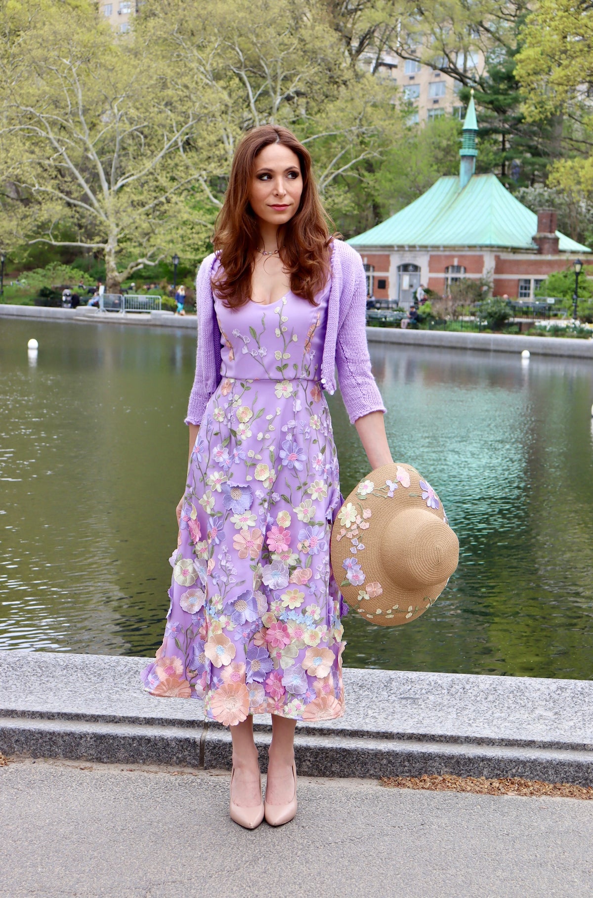 Model standing near pond in lilac midi length satin appliquéd floral details with matching lilac cropped knitted lilac sweater and matching floral appliqued straw sun hat in hand to show full aerial of hat.