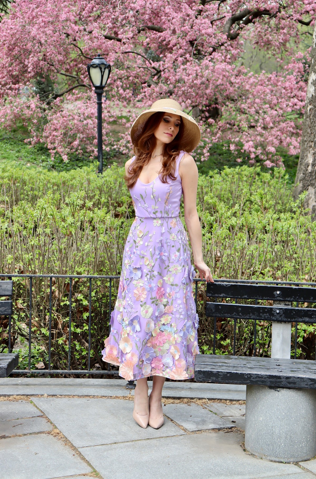 Model standing near park bench in lilac midi length appliquéd floral dress with floral applique detail straw sun hat.