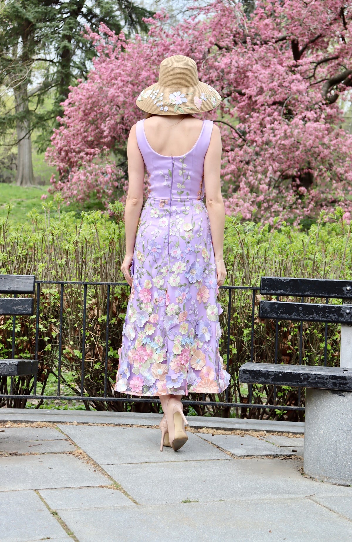 Back view of Model  to show off back of lilac midi length satin floral appliquéd dress with blush, lilac, and yellow florals with straw sun hat with floral applique details