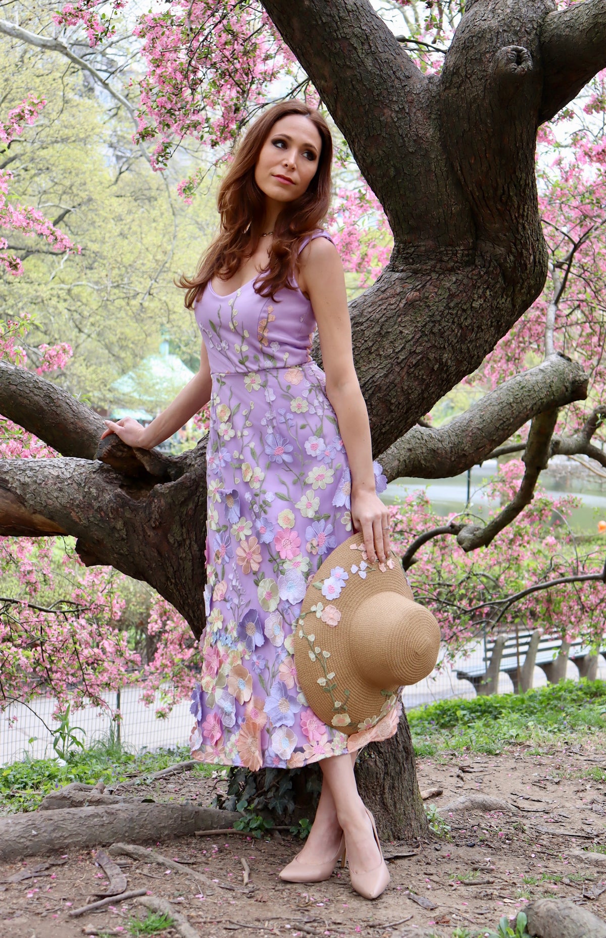 Model leaning on cherry blossom tree with sleeveless lilac midi length floral appliquéd dress with straw sun hat with floral appliques in hand to show aerial view of hat.