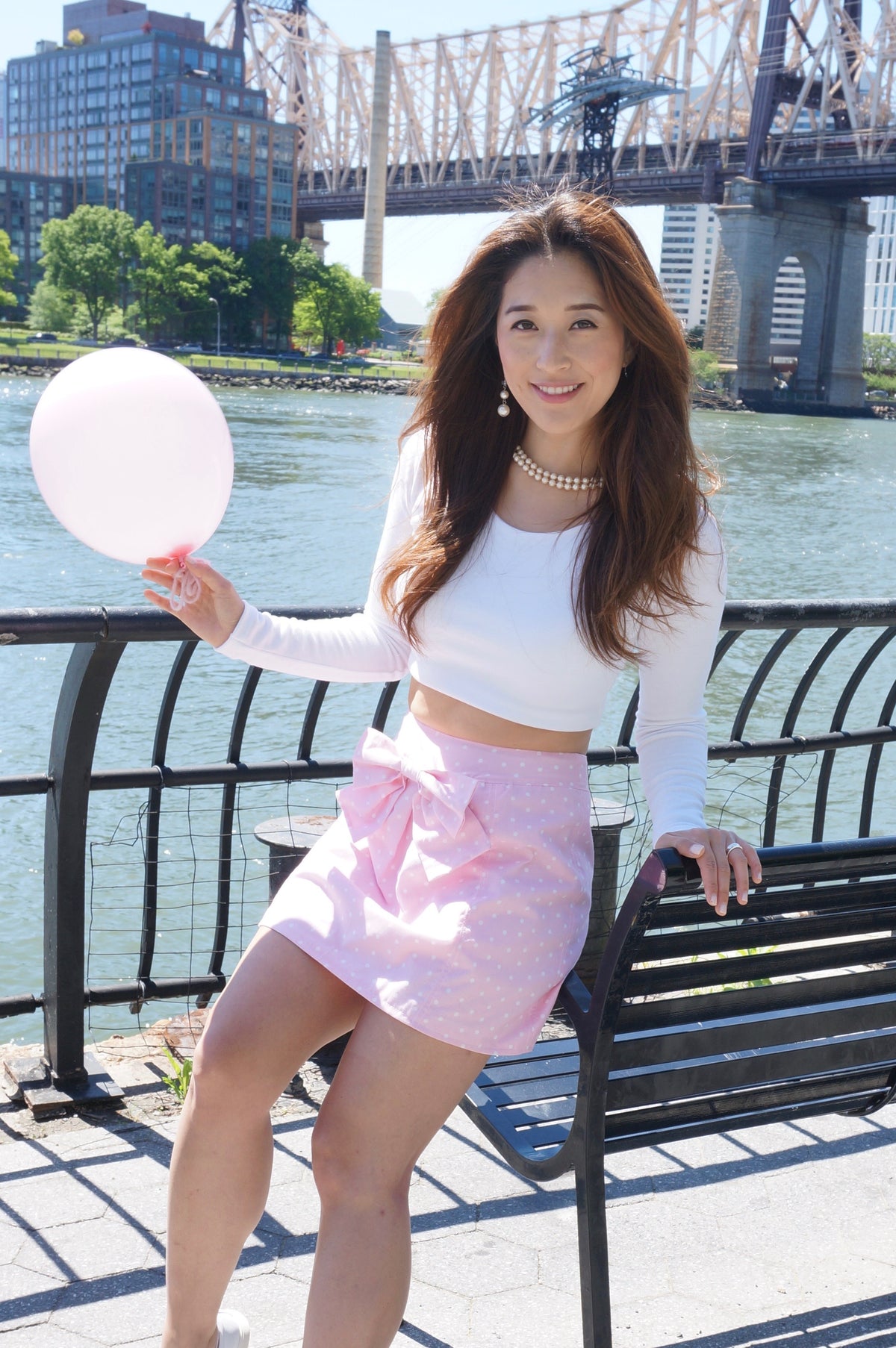Model wearing a white long sleeve jersey knit crop top and a pink and white polka dot print skirt with front bow sitting on a bench holding a pink balloon smiling.