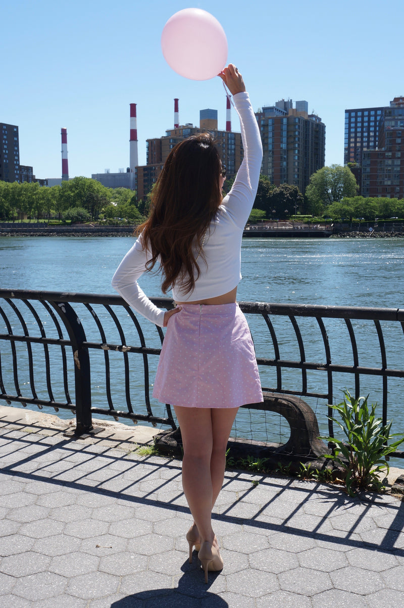 Back of model wearing a white long sleeve jersey knit crop top and a pink and white polka dot print skirt with one hand on her hip and the other in the air holding a balloon.