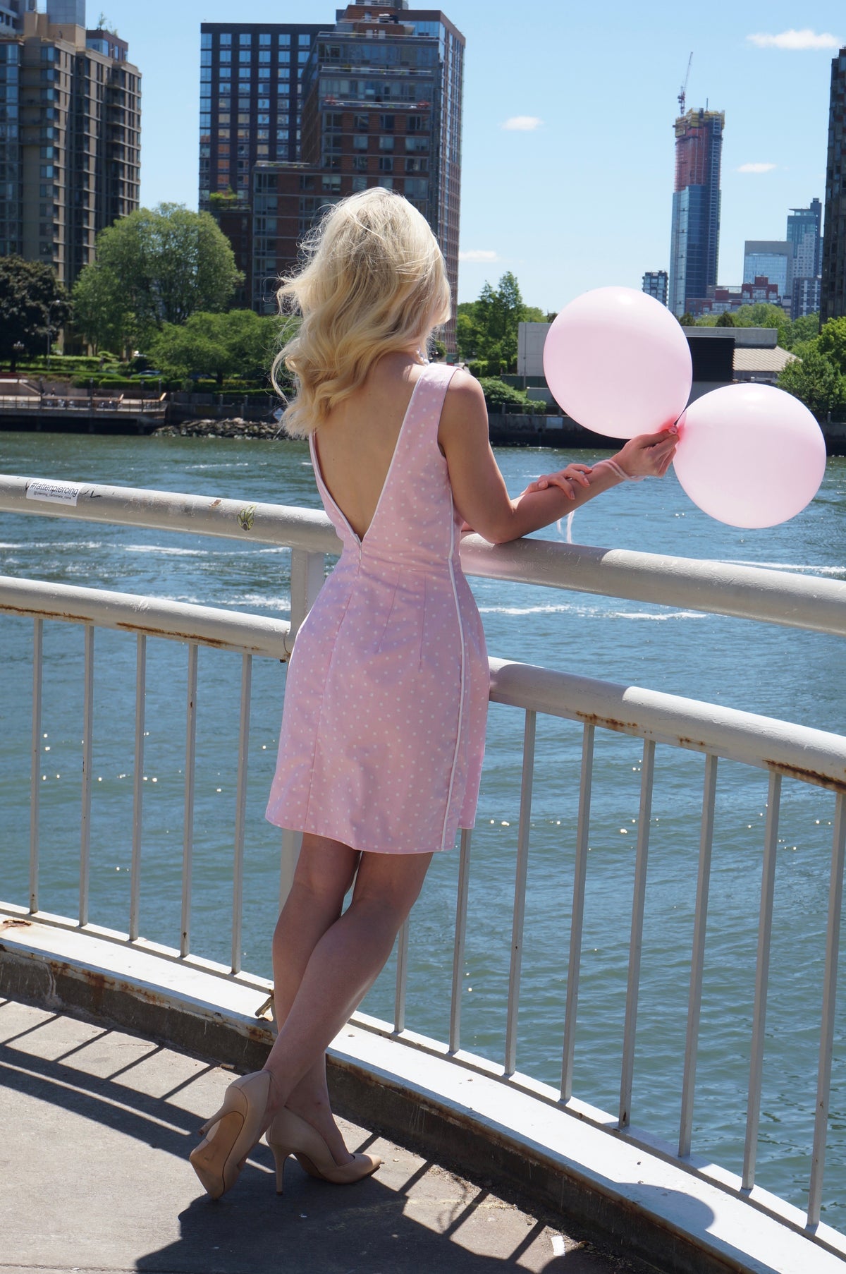 Back of model wearing pink and white polka dot print dress with white piping holding two balloons in front of water.