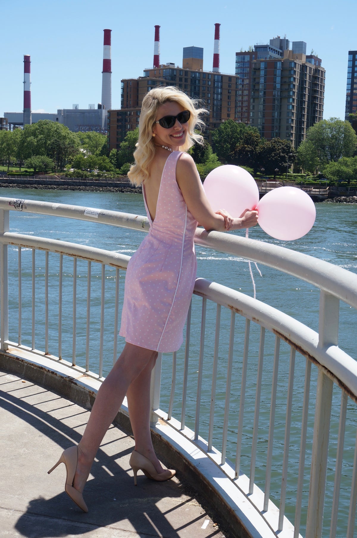 Side view of model wearing pink and white polka dot print dress with white piping and low back smiling over her shoulder, holding two pink balloons.