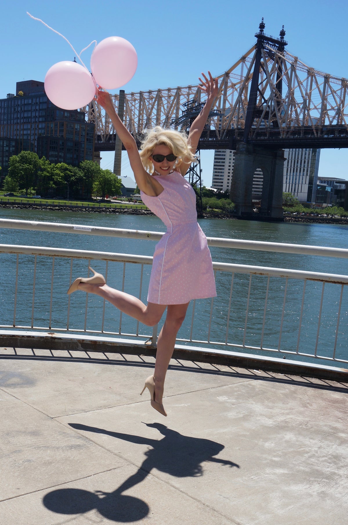 Side view of model wearing pink and white polka dot print dress with white piping smiling as she jumps in the air holding two pink balloons. 