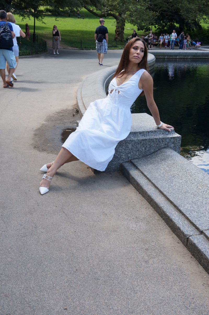 Model wearing a white floral eyelet fabric and a low V-neck with a bow top sitting in front of a pond.