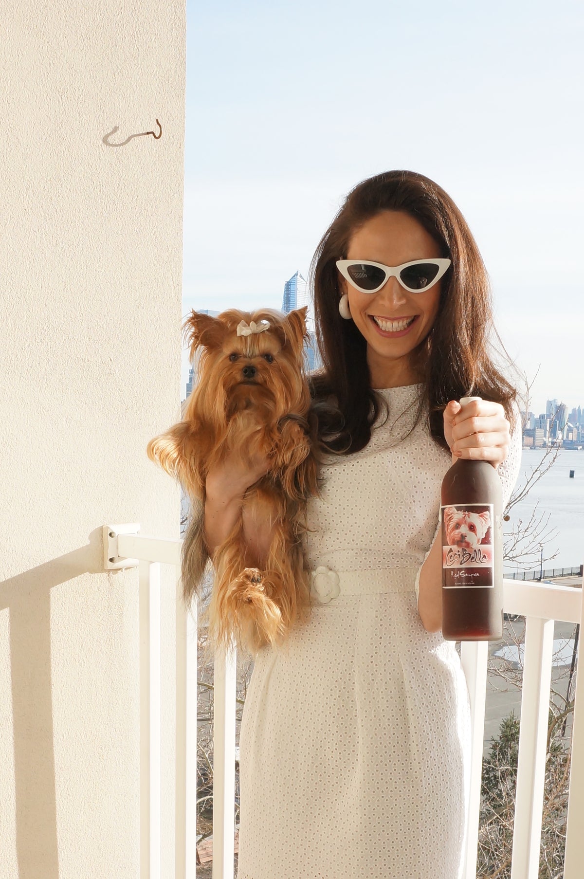 Model wearing a 100% cotton eyelet, fully lined cotton dress, with peekaboo sleeves, and a belt with a flower buckle smiling on a balcony in front of the city holding a Yorkie and a bottle of Wine.