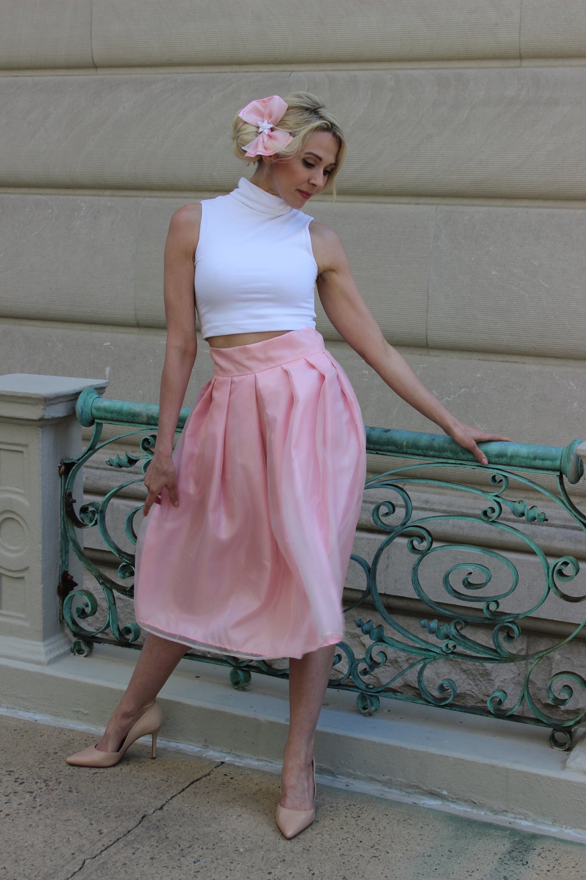 Model wearing a full satin and organza skirt atop a layer of tulle netting and a white turtleneck crop top and a pink bow in her hair.