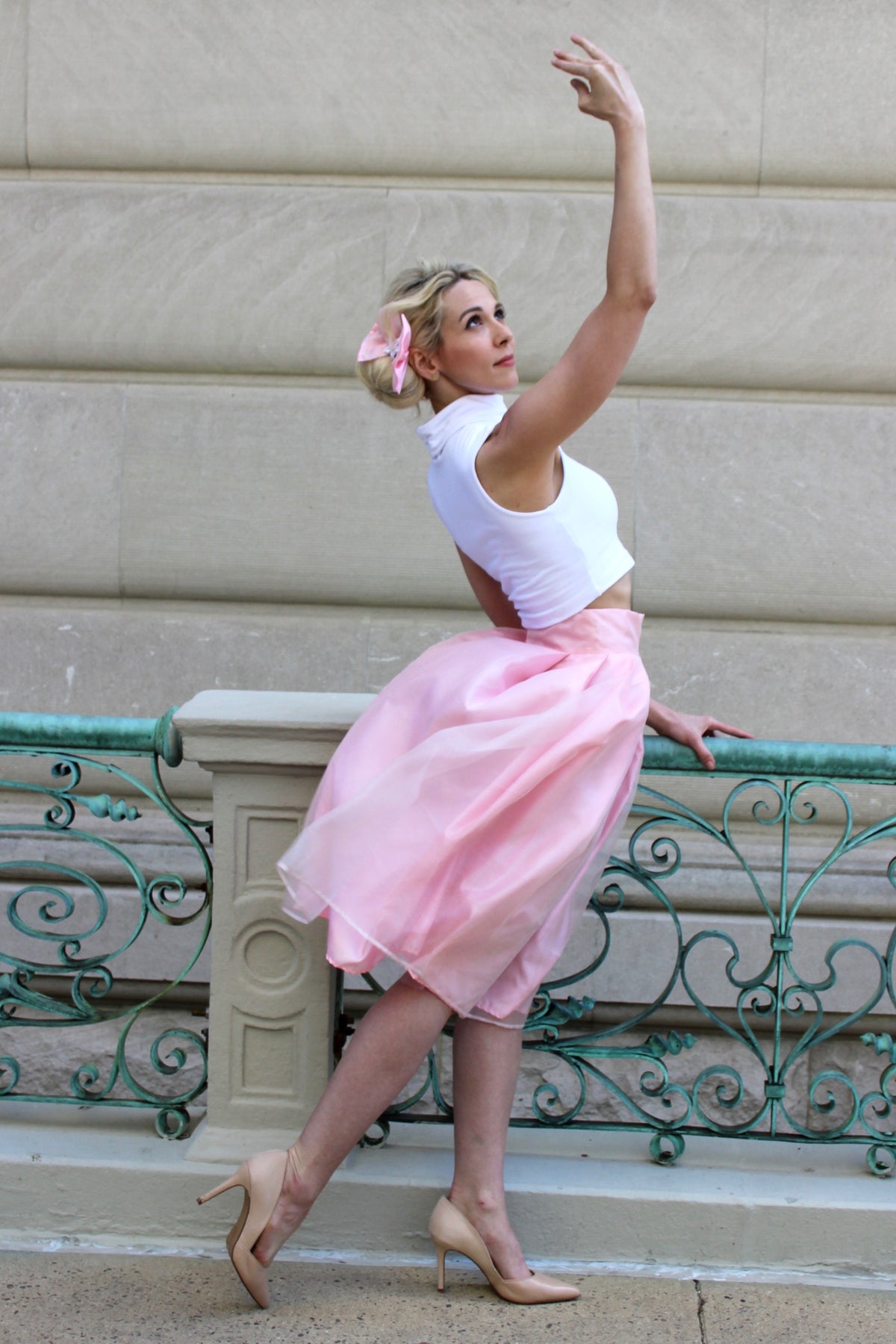 Model wearing a full satin and organza skirt atop a layer of tulle netting and a white turtleneck crop top and a pink bow in her hair in a classic ballet position.