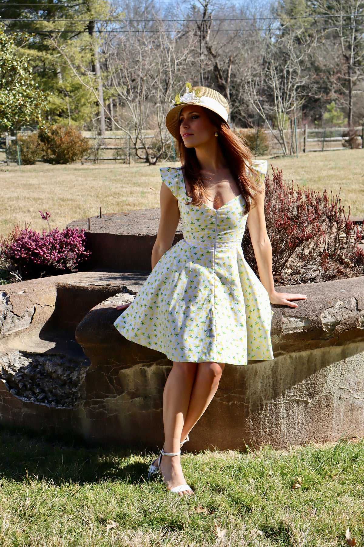 Model in lemon print dress wearing a straw hat sitting on a rock, looking into the distance.