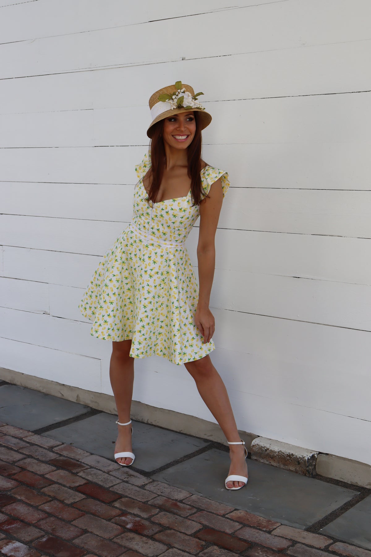 Model in lemon print dress wearing a straw hat in front of a white wall, smiling and looking off to the side.