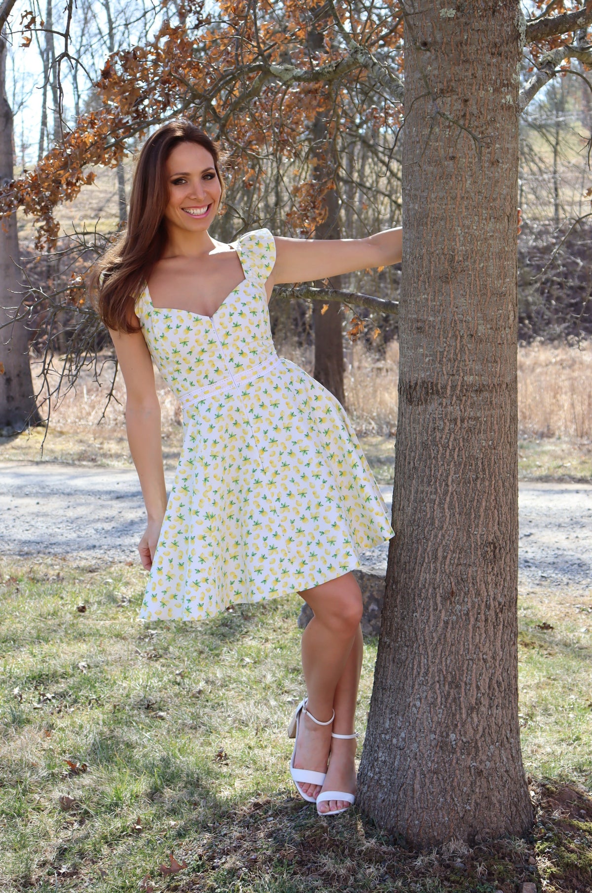 Model in lemon print dress posing next to a tree, smiling.