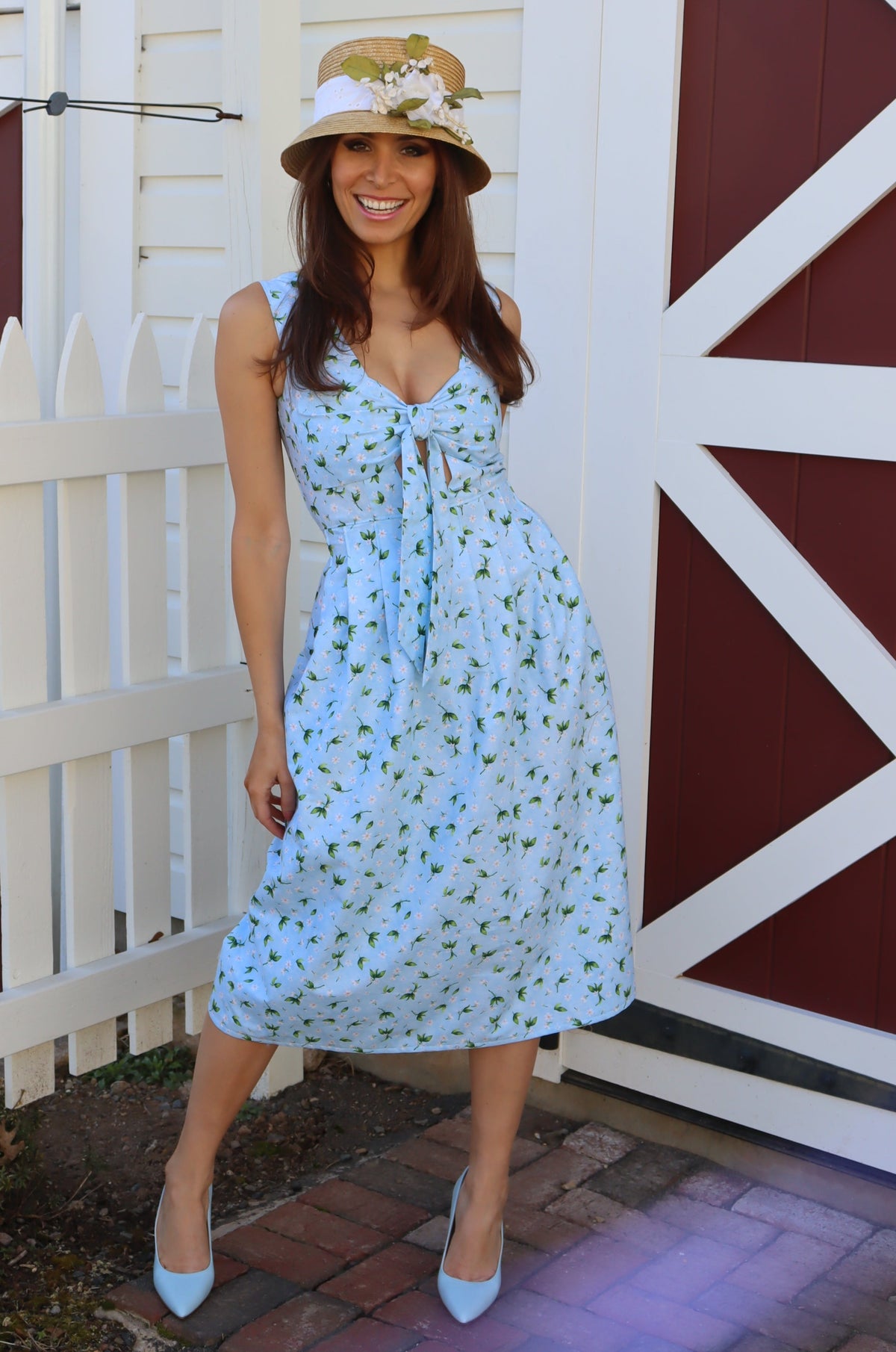 Model in wicker hat in front of barn, wearing a midi light blue dress with peek-a-boo bow front, in front of a barn.