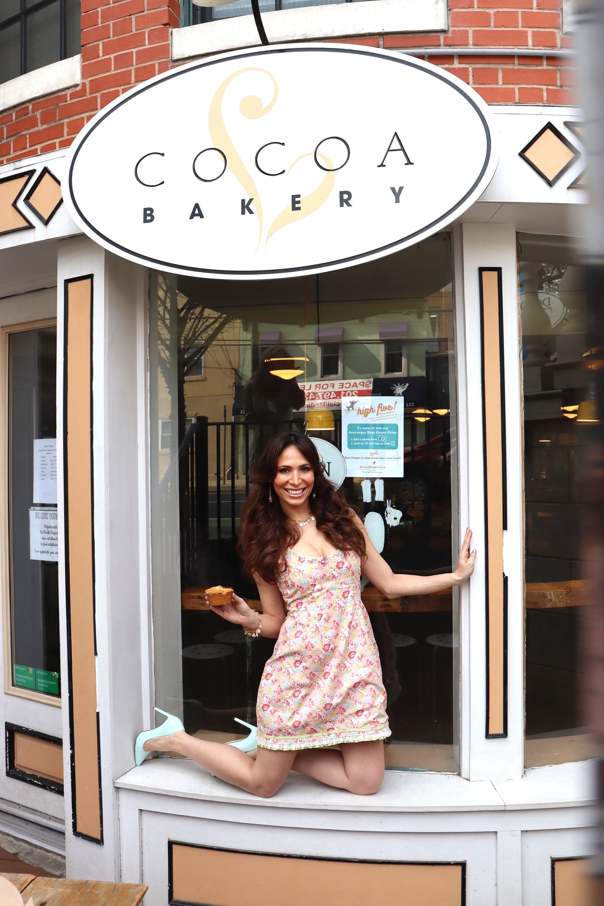 Model wearing a pink, green and blue floral print dress smiling in the window of Cocoa Bakery.