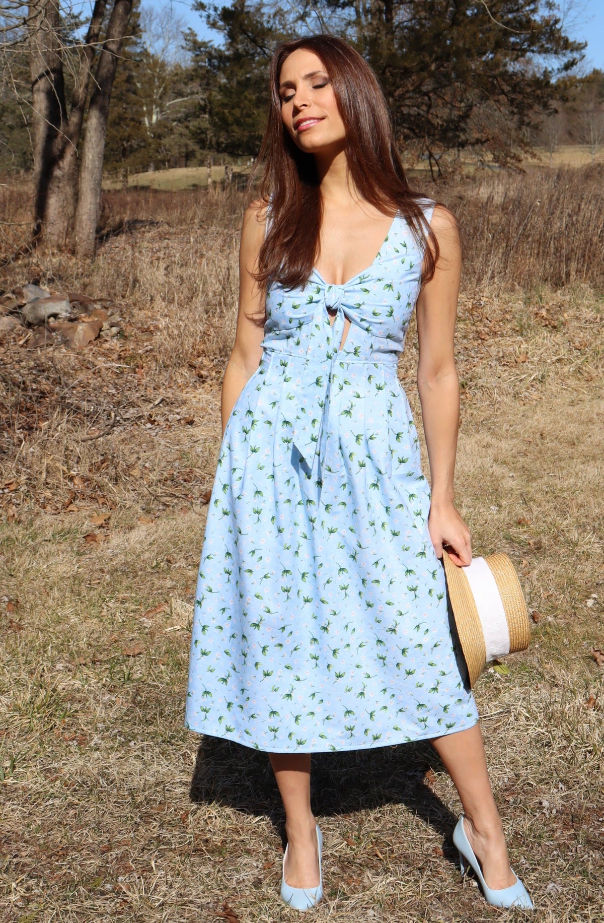 Model wearing a midi light blue dress with peek-a-boo bow front, on wintered grass.