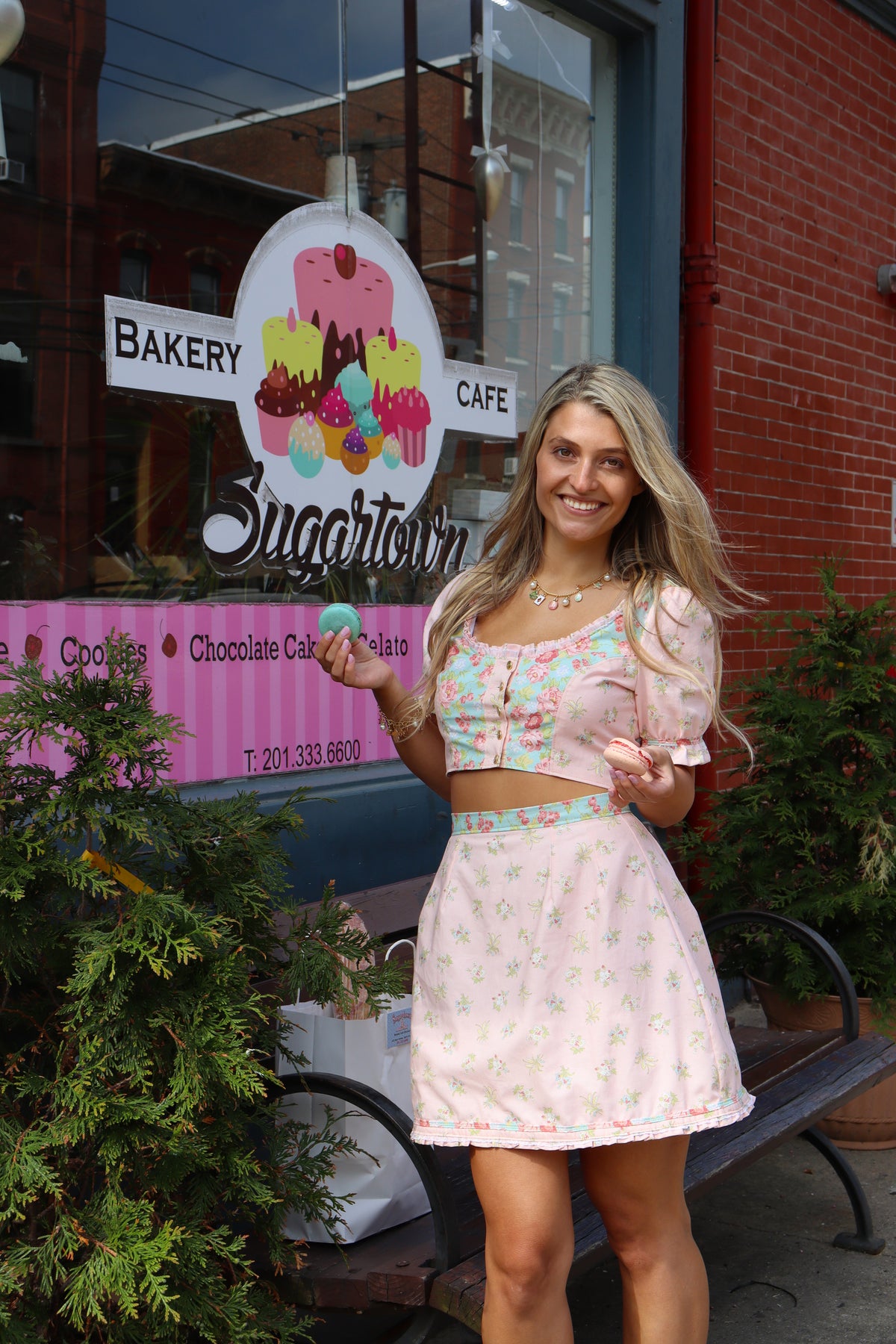 Model wearing a pink and aqua, 3/4 sleeve crop top with gold stitching and a pink and aqua skirt with gold top stitching smiling and holding one blue macaron in one hand and one pink macaron in the other.