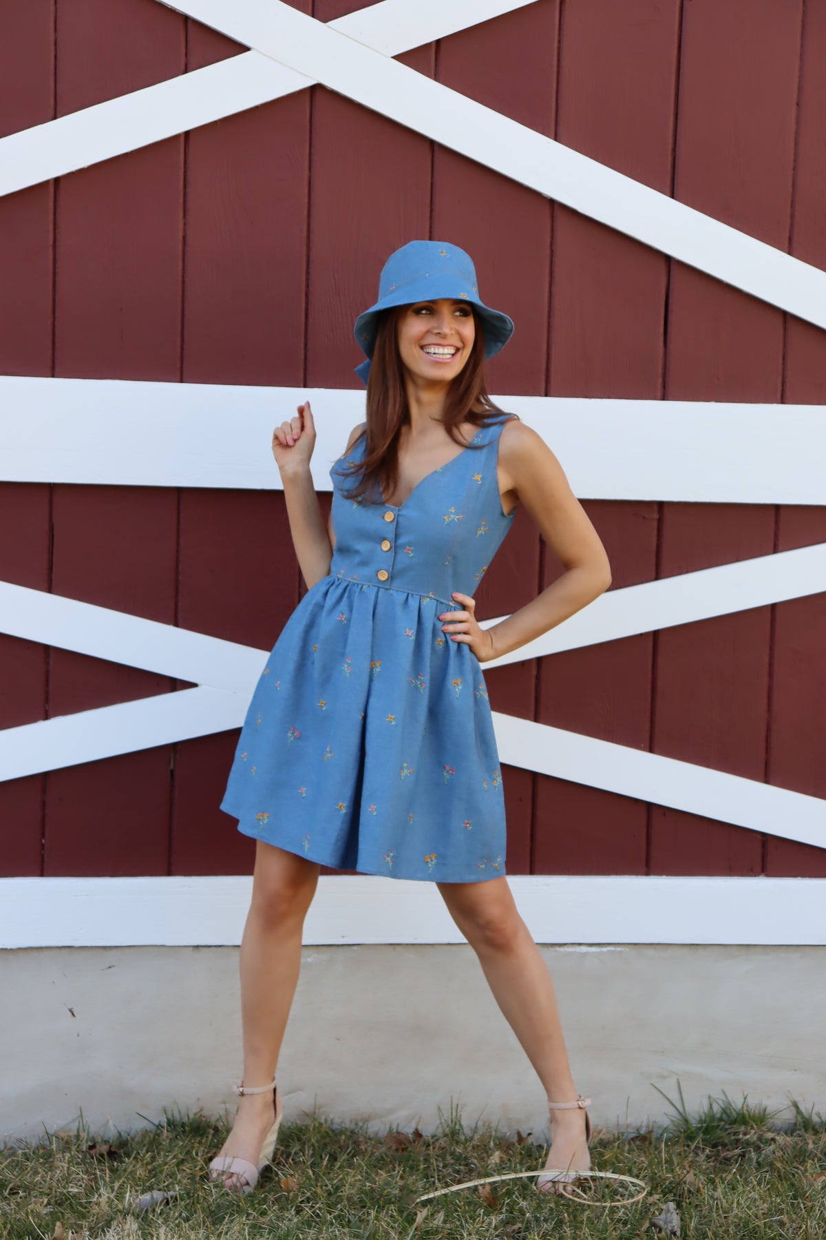 Model in floral embroidered denim dress wearing a floral embroidered denim bucket hat with bow in front of a barn, smiling with one hand at her hip and the other angling upwards with elbow tucked at her hip.
