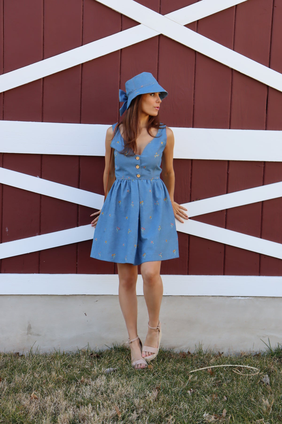 Model in floral embroidered denim dress wearing a floral embroidered denim bucket hat with bow, looking off to the side with hands reaching back against a barn.