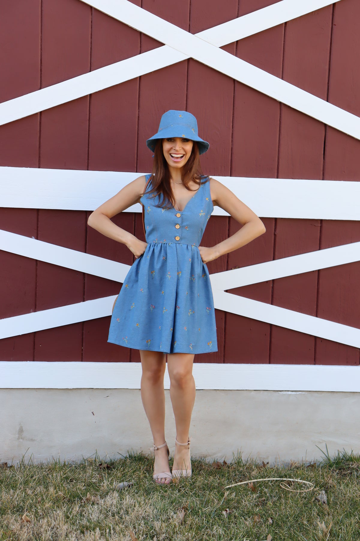 Model in floral embroidered denim dress wearing a floral embroidered denim bucket hat with bow in front of a barn, smiling with hands on her hips.