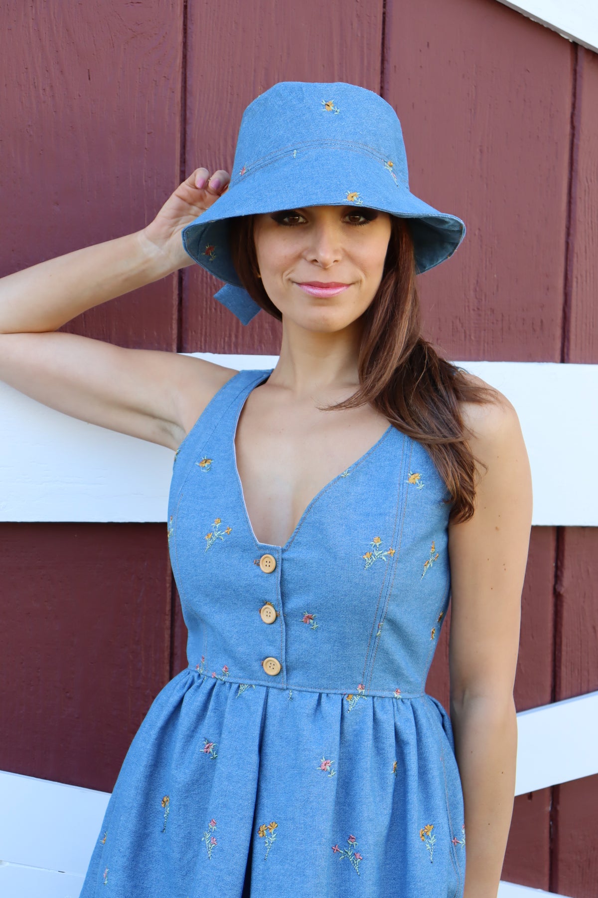 Close up of model in a floral embroidered denim dress wearing a floral embroidered denim bucket hat with bow in front of a barn with arm bent towards head, smiling softly.