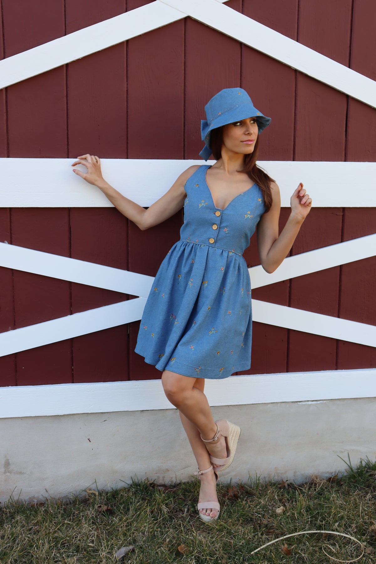 Model in floral embroidered denim dress wearing a floral embroidered denim bucket hat with bow in front of a barn, with one arm bent towards the barn, the other angling upwards, looking off to the side.