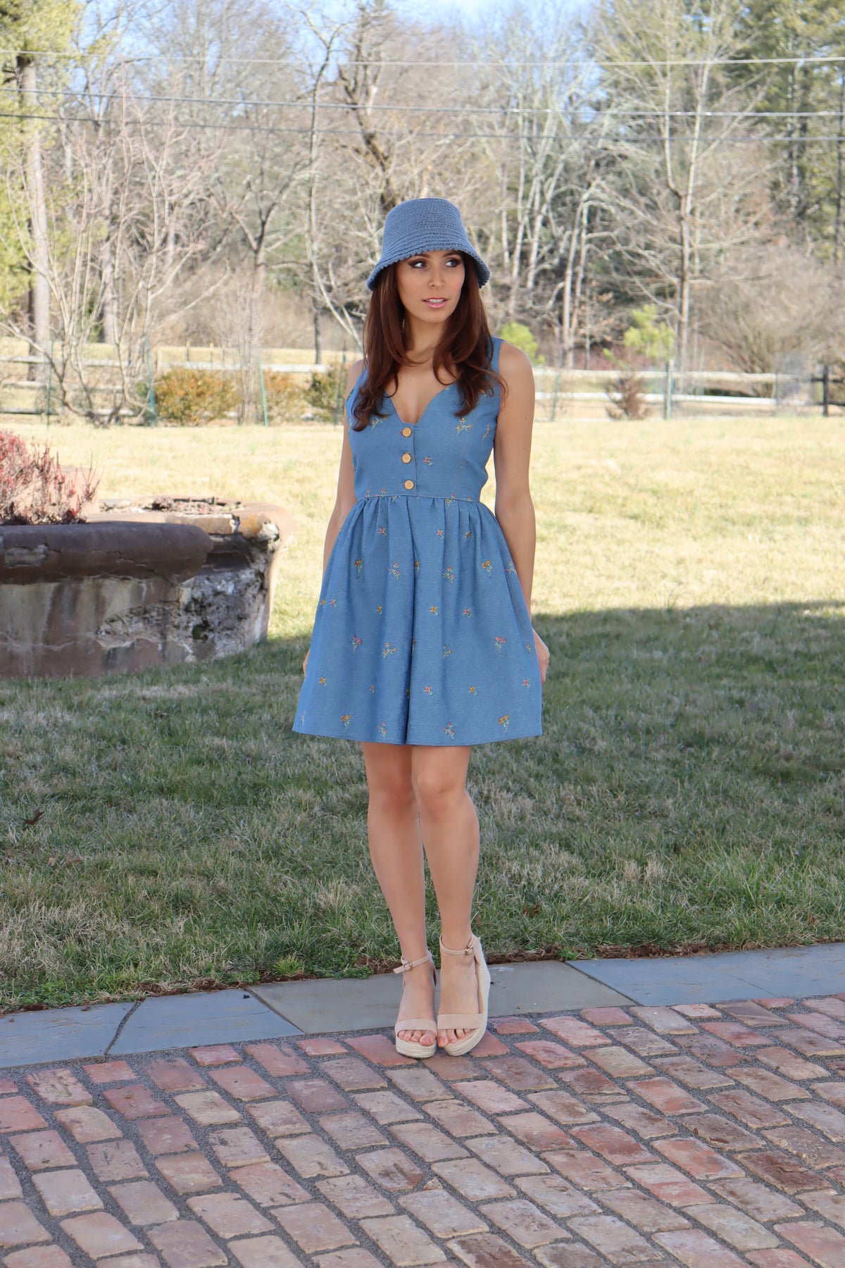 Model in floral embroidered denim dress wearing a hand crocheted blue bucket hat with bow in front of a field of grass, looking off to the side.