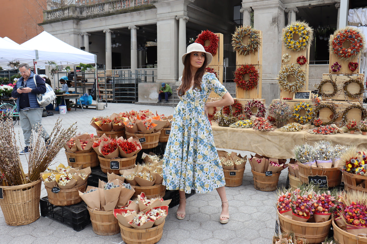 Model wearing midi length dress of a yellow and blue floral print with arms behind her in front of a dried floral display.