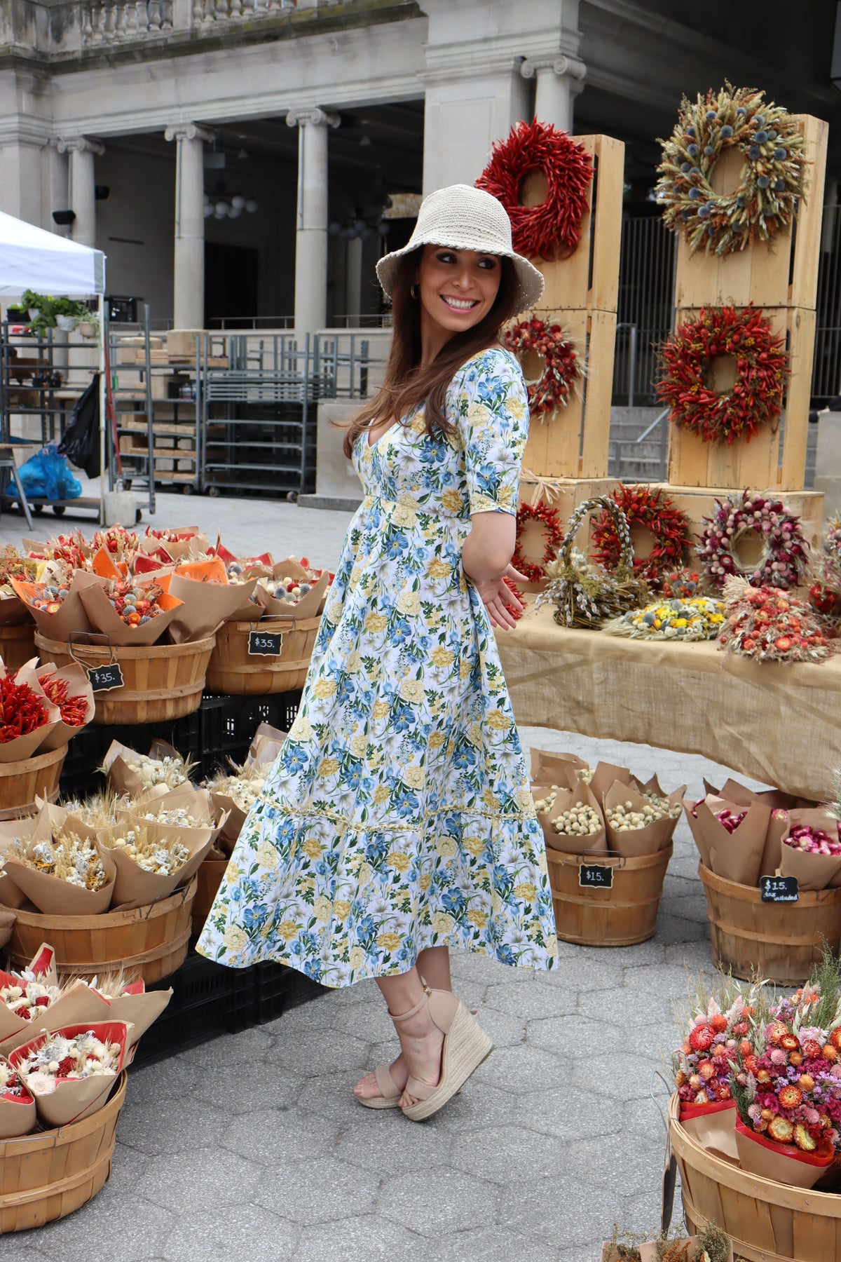 Side view of Model wearing midi length dress of a yellow and blue floral print with arms behind her in front of a dried floral display.