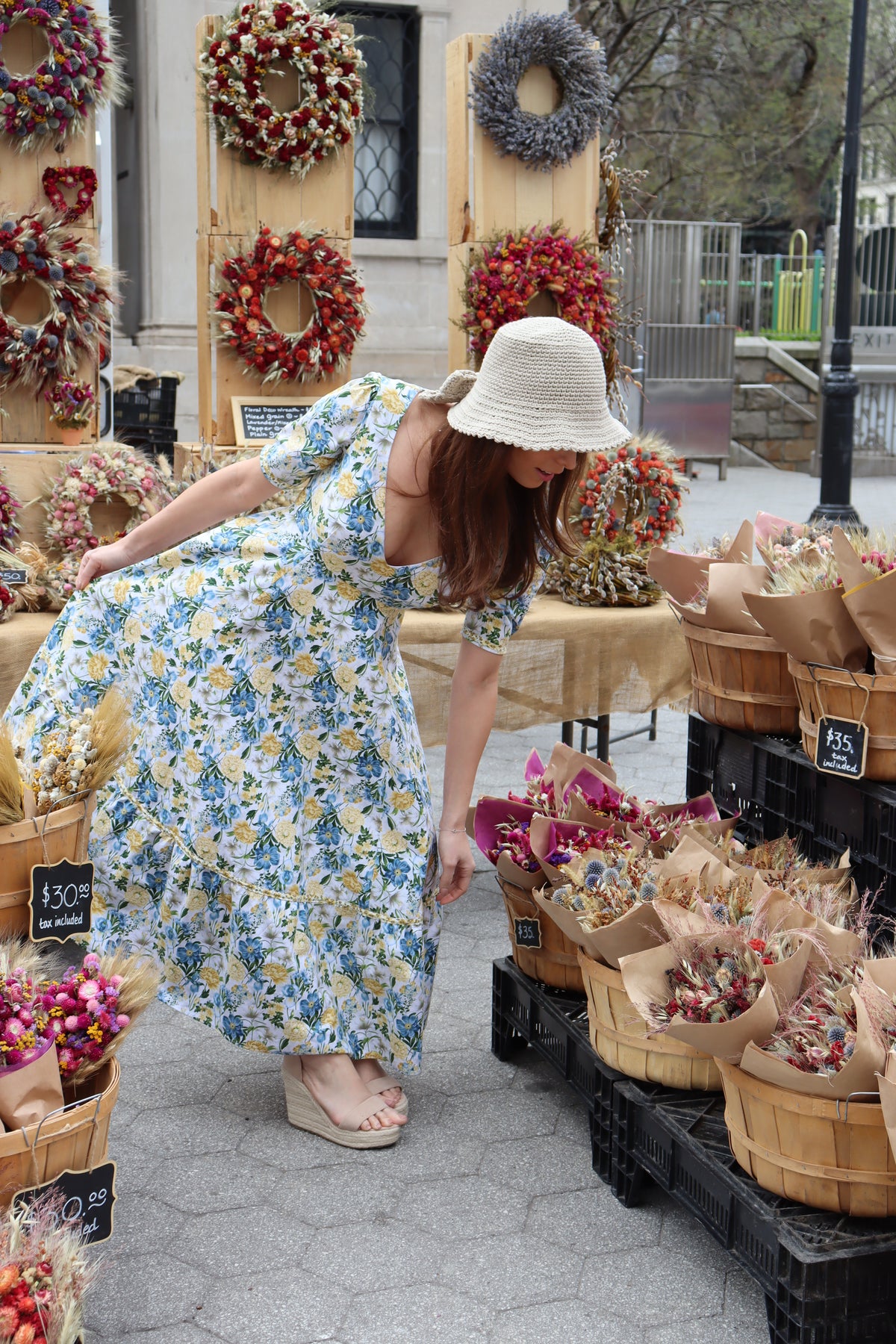 Model bending down wearing midi length dress of a yellow and blue floral print in front of a dried floral display.