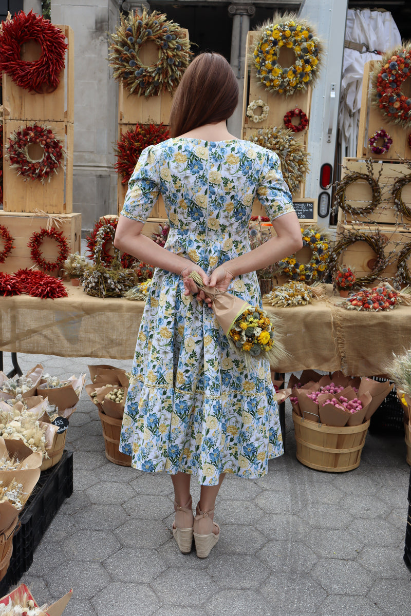 Back view of Model wearing midi length dress of a yellow and blue floral print in front of a dried floral display.