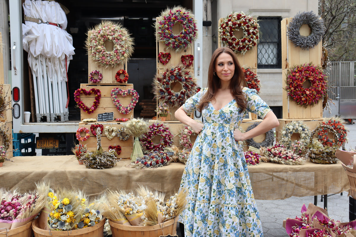 Model wearing midi length dress of a yellow and blue floral print with hands on hips in front of a dried floral display.