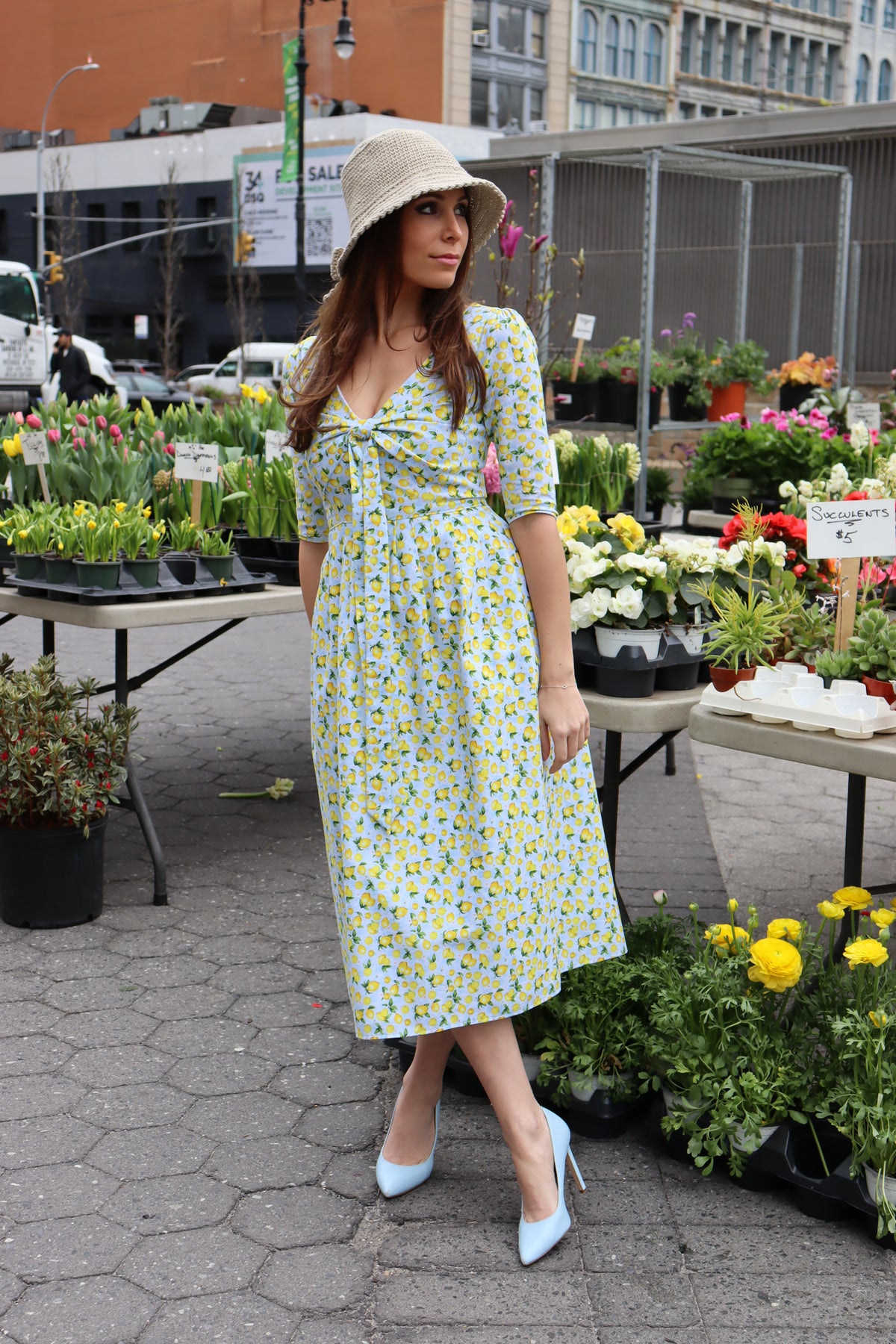 Model wearing a tan bucket hat and a midi dress in a lemon print of aqua blue and  yellow in front of a flower display at a farmer's market.