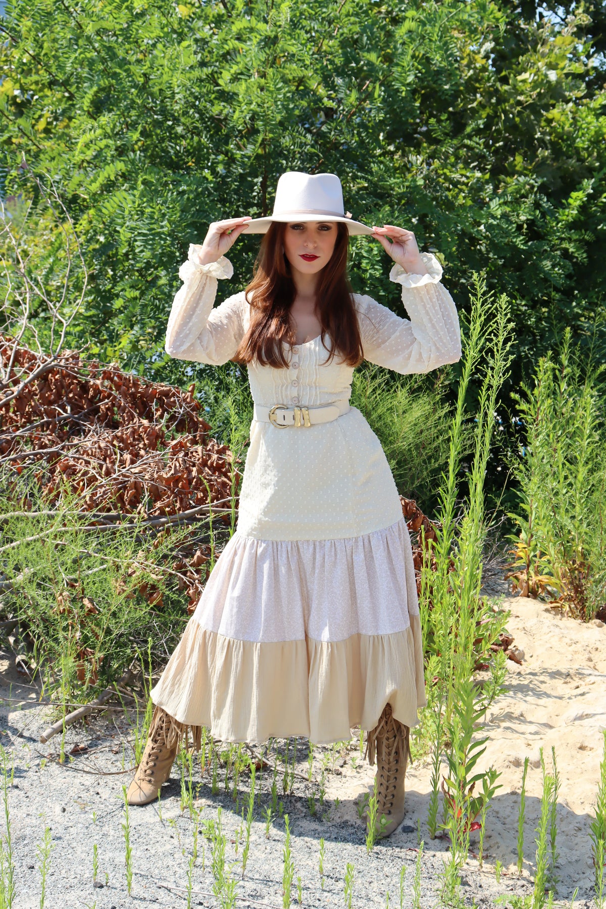 Model wearing dress with three different fabrics, Swiss Dot, Calico and Gauze midi dress, a belt, and hat with both hands touching the brim.