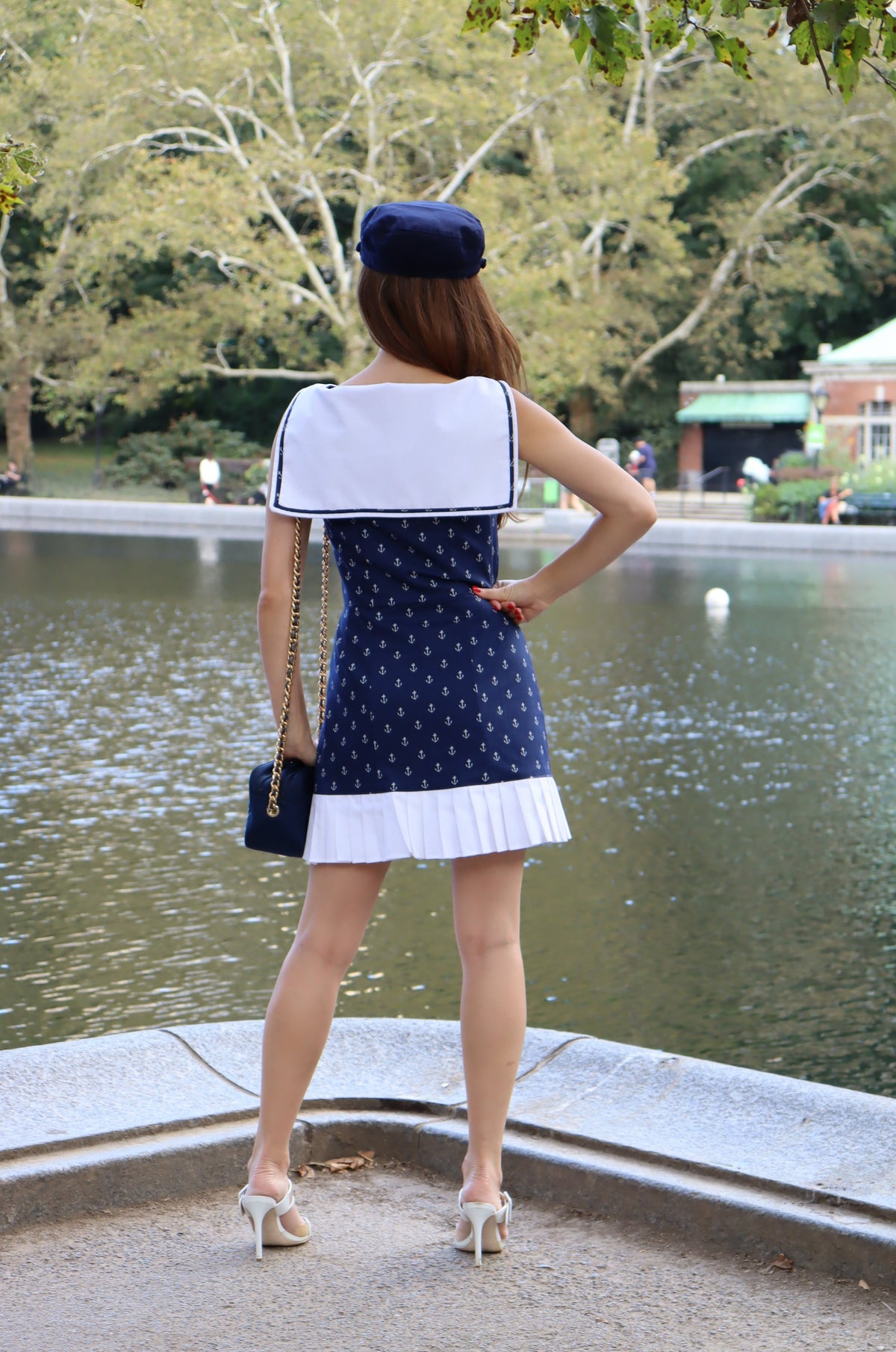 Back of model wearing classic sailor dress with white anchors on navy background print with ruffle at the bottom and a red bow and a blue hat with her hand on her hip.