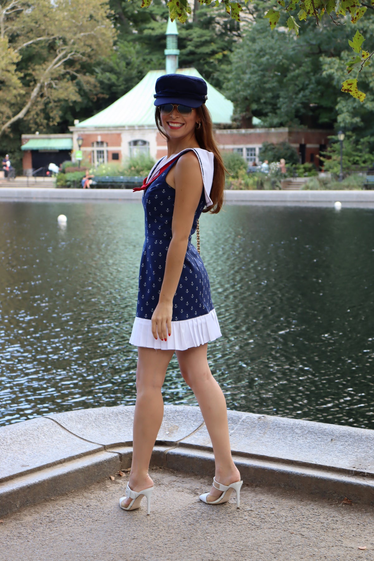 Model wearing classic sailor dress with white anchors on navy background print with ruffle at the bottom and a red bow, and a blue hat smiling.