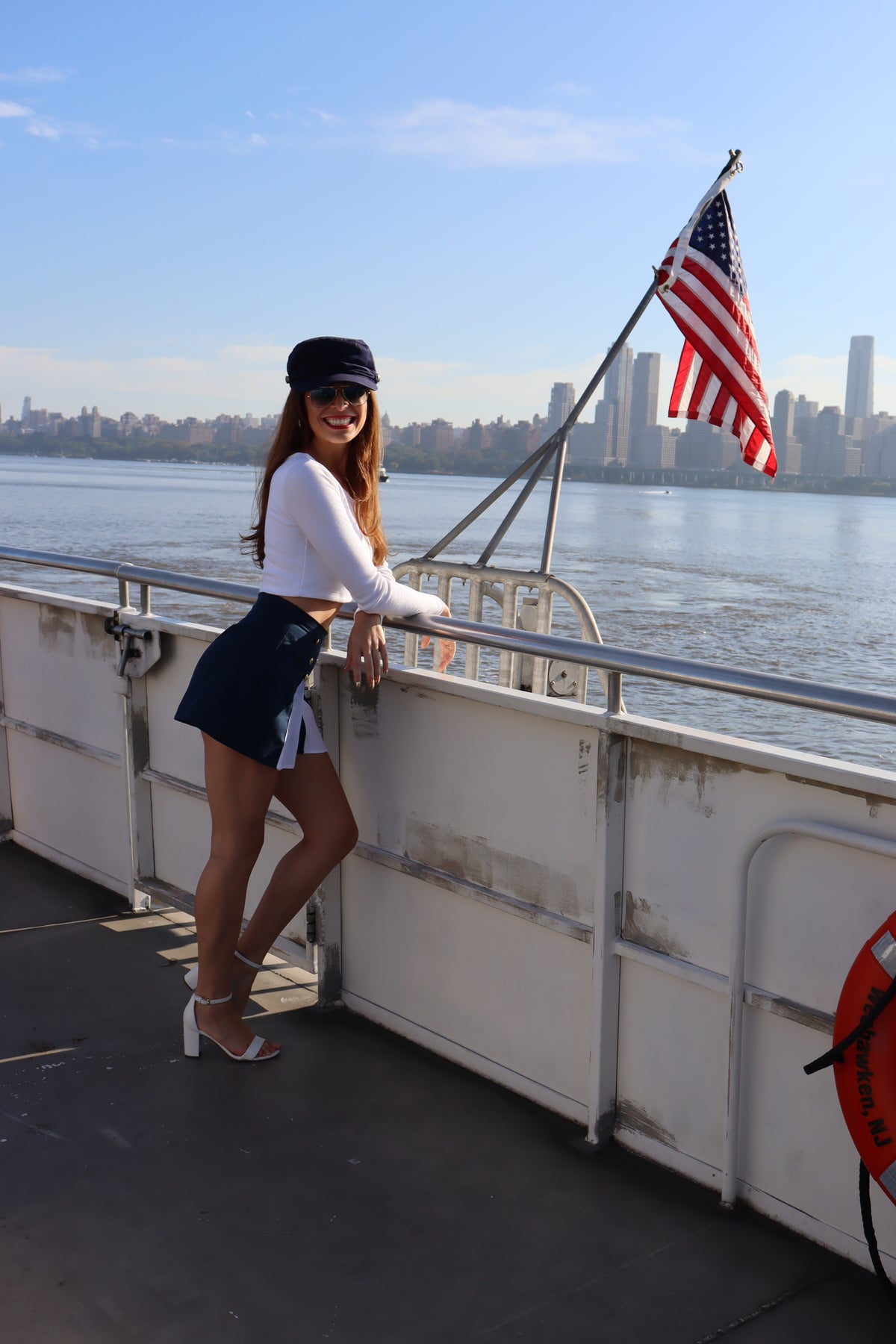 Model wearing scoop neck long sleeve white crop top and blue and white spinnaker shorts leaning against a railing in front of the American flag in front of water.