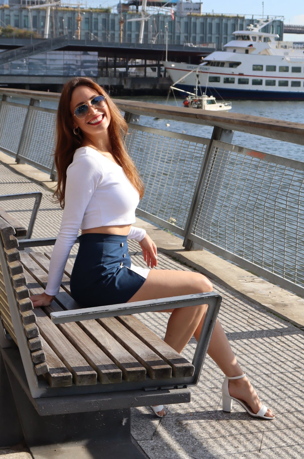 Model wearing scoop neck long sleeve white crop top and blue and white spinnaker shorts sitting down on a bench smiling.