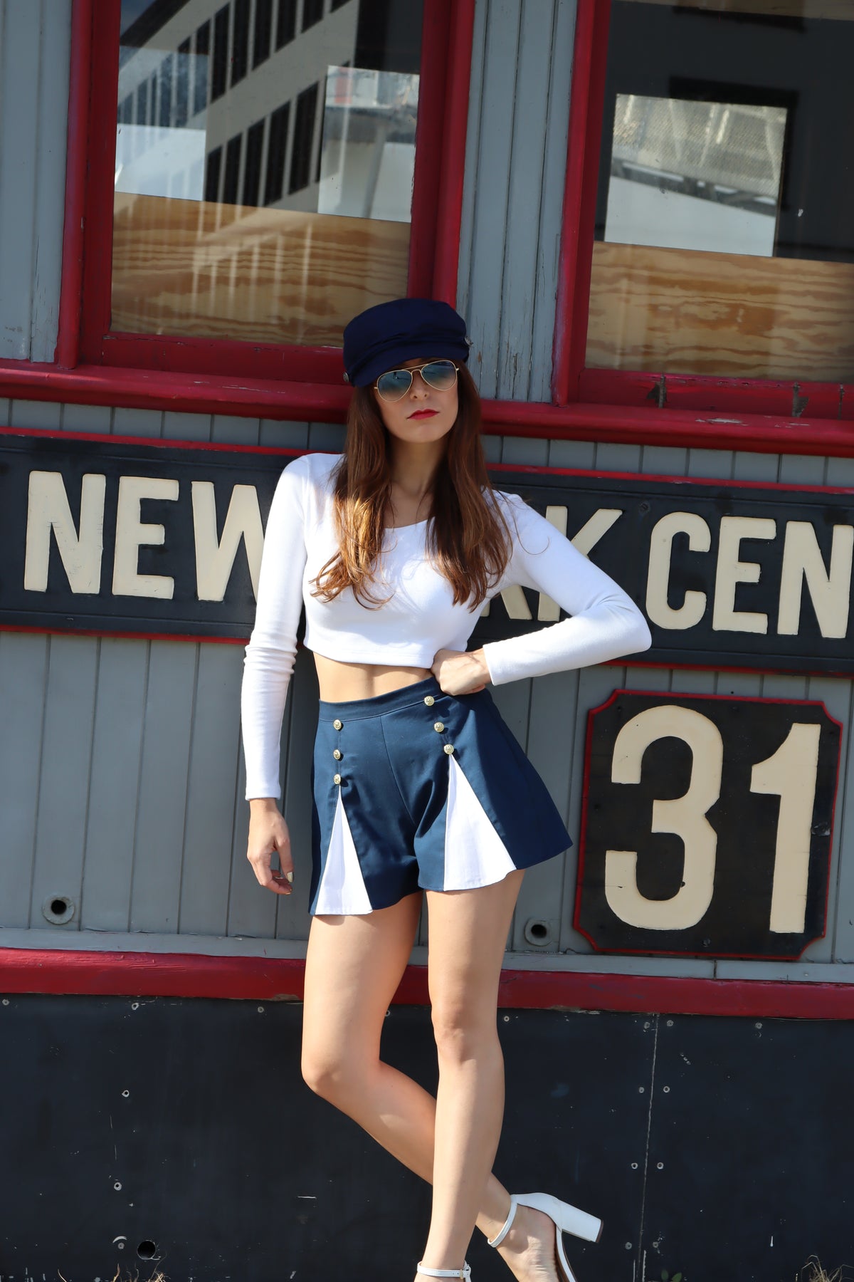 Model wearing scoop neck long sleeve white crop top, blue and white spinnaker shorts with gold buttons, and blue hat in front of sign.