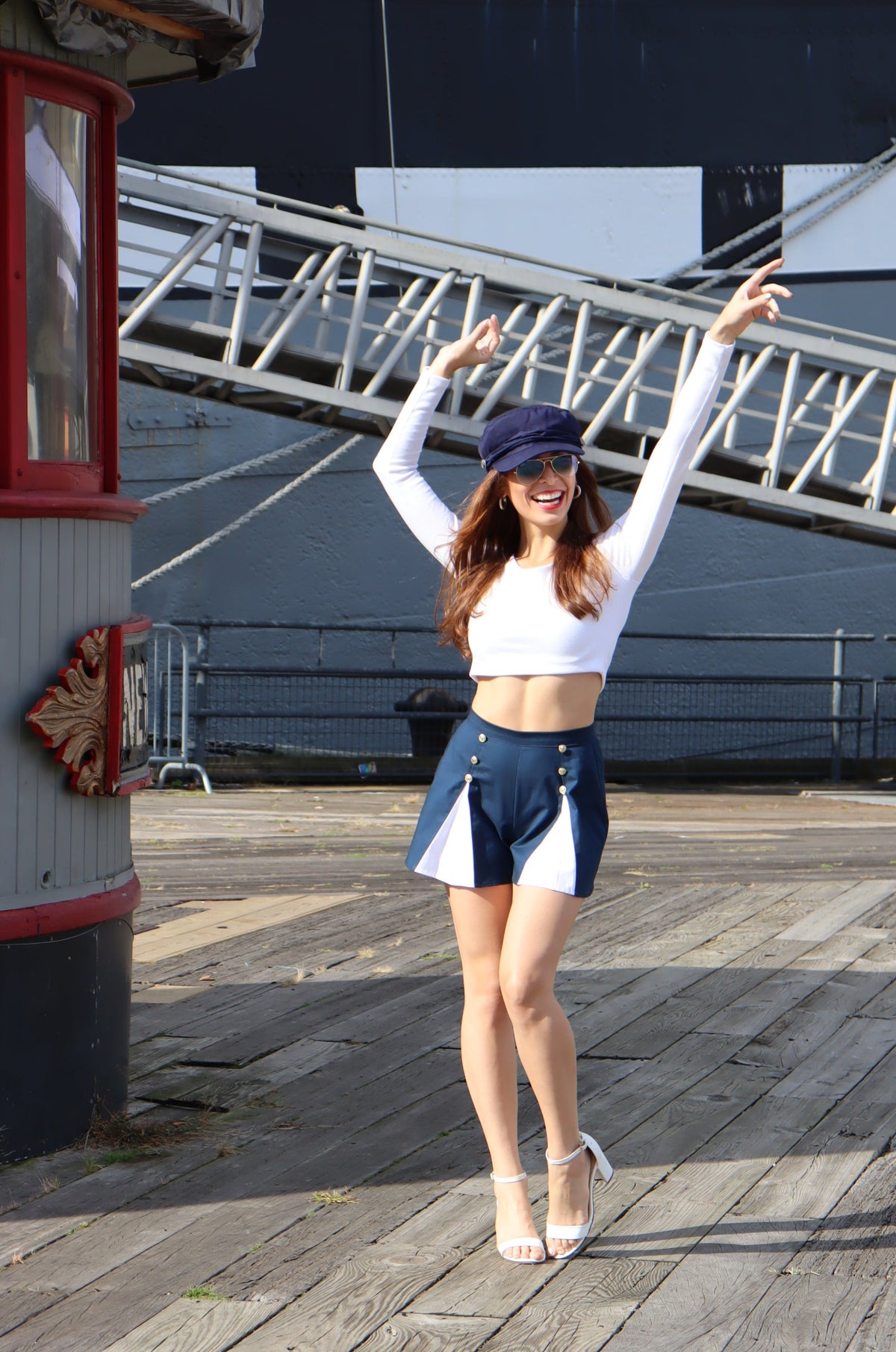 Model wearing scoop neck long sleeve white crop top and blue and white spinnaker shorts with her arms in the air smiling.