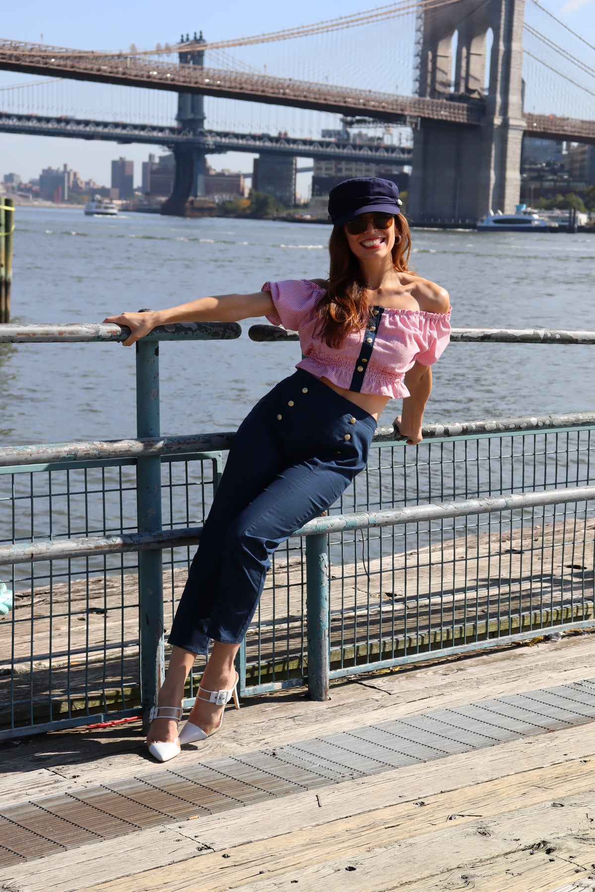 Model wearing jib top, navy cotton port capris and blue hat leaning against a railing smiling.