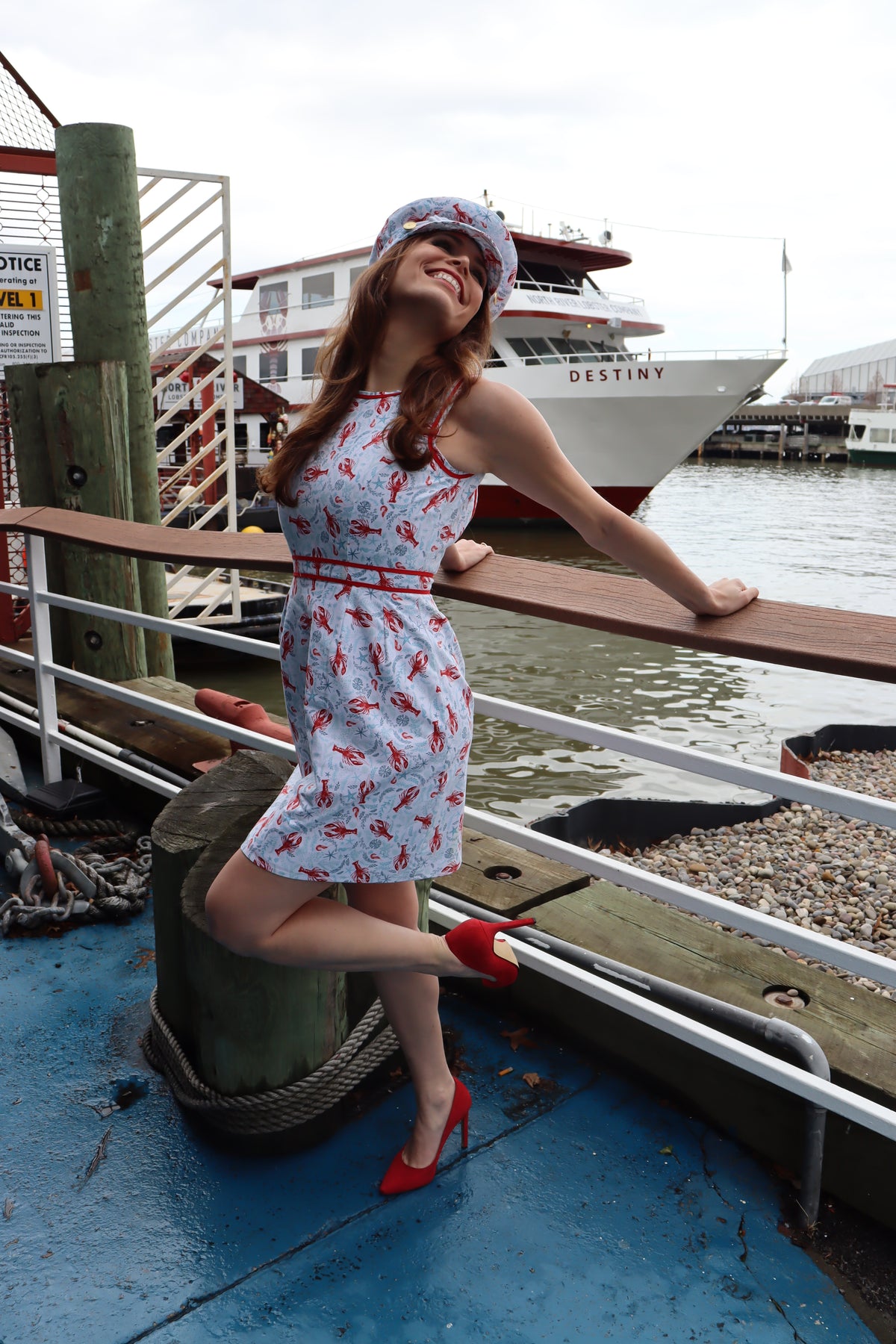 Model wearing light blue and red lobster on white background print dress with matching hat smiling reaching back towards railing in front of a red and white boat.