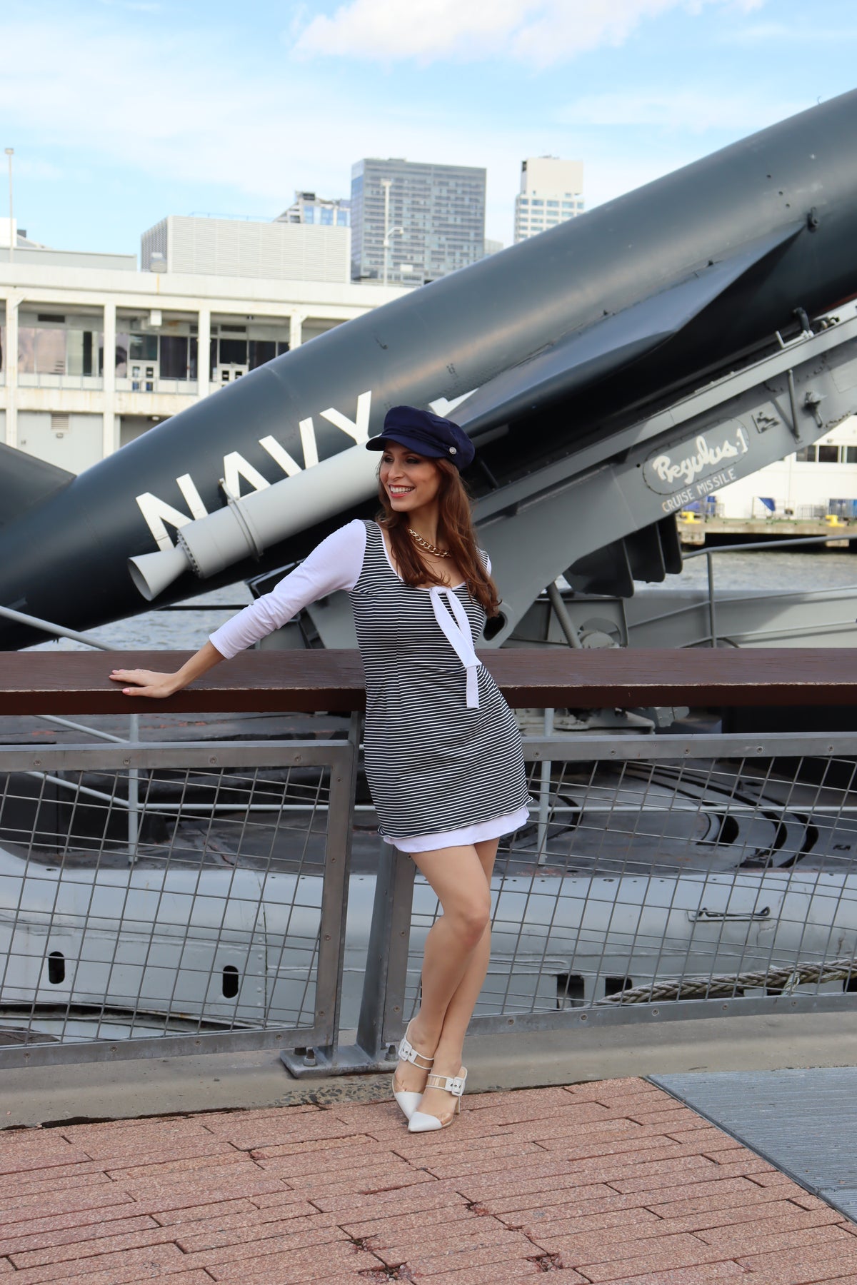 Model wearing navy and white pinstripe on top with white underneath t-shirt dress and blue hat in front of a blue navy plane smiling.