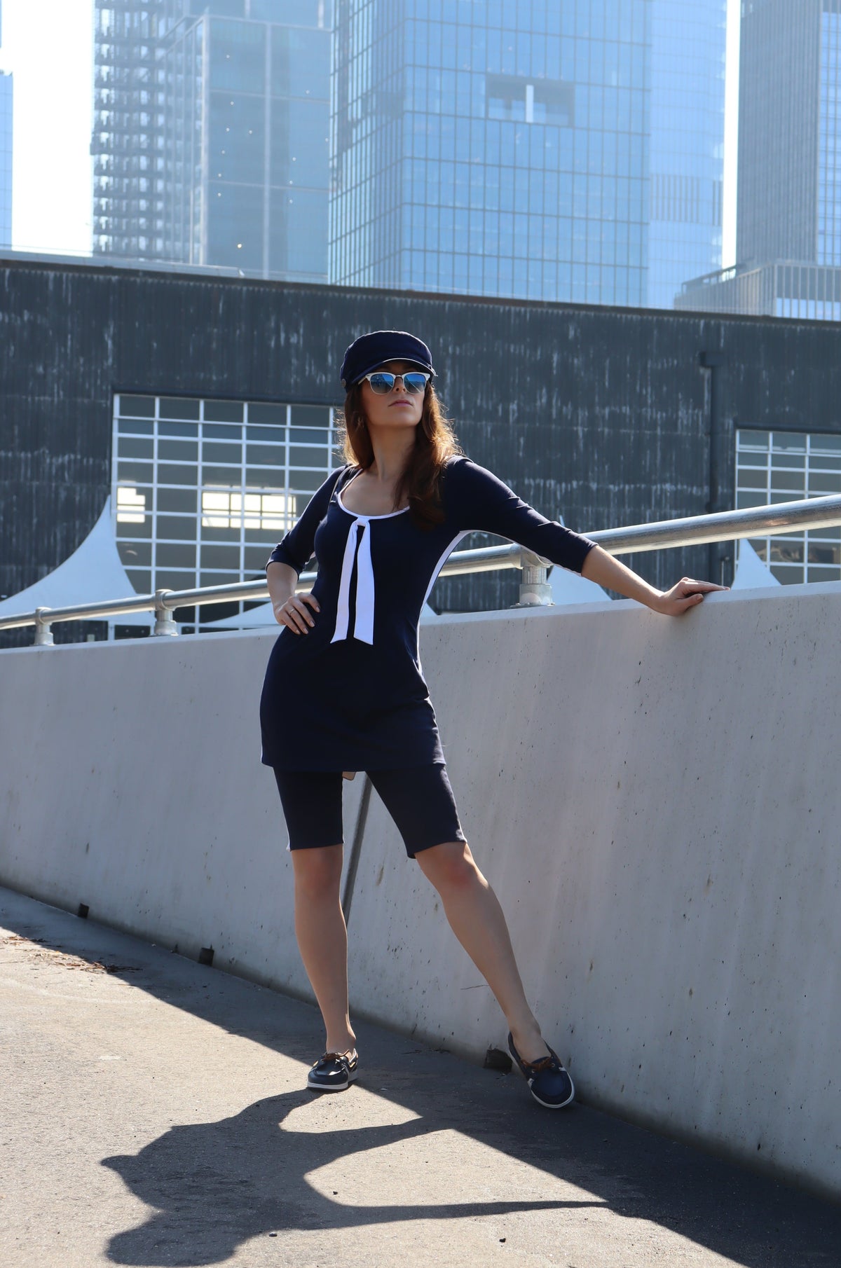 Model wearing navy with white stripes down the sides, 3/4 sleeves and a white tie tunic and a blue hat with one hand on her hip.